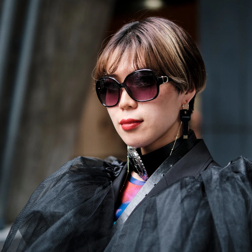 woman in a black jacket with puffy sleeves, sunglass, and a red lip, with a pixie haircut and bangs