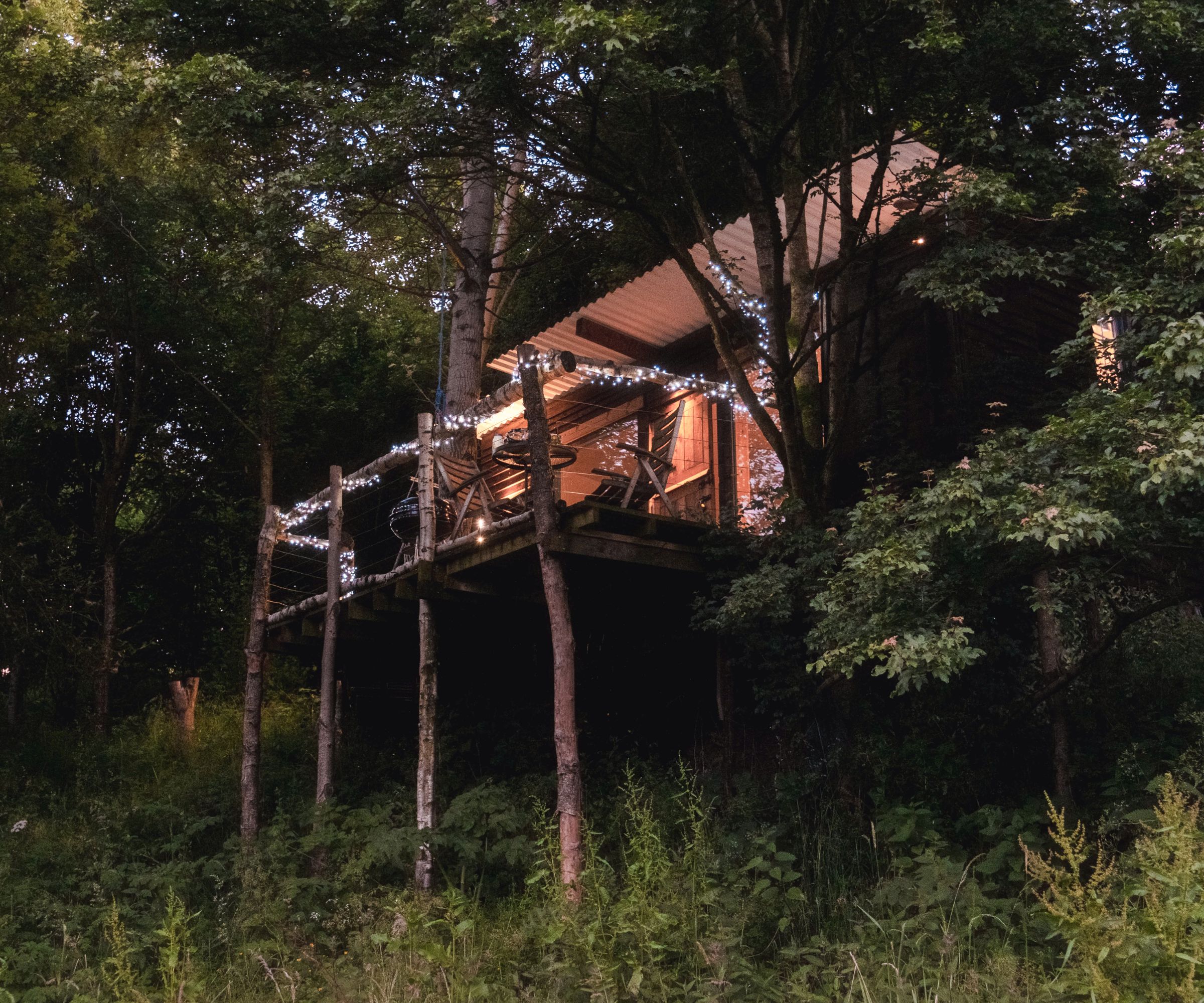 A wooden treehouse with a covered deck, decorated with string lights, set on tall stilts among dense forest greenery and softly lit from within at dusk.