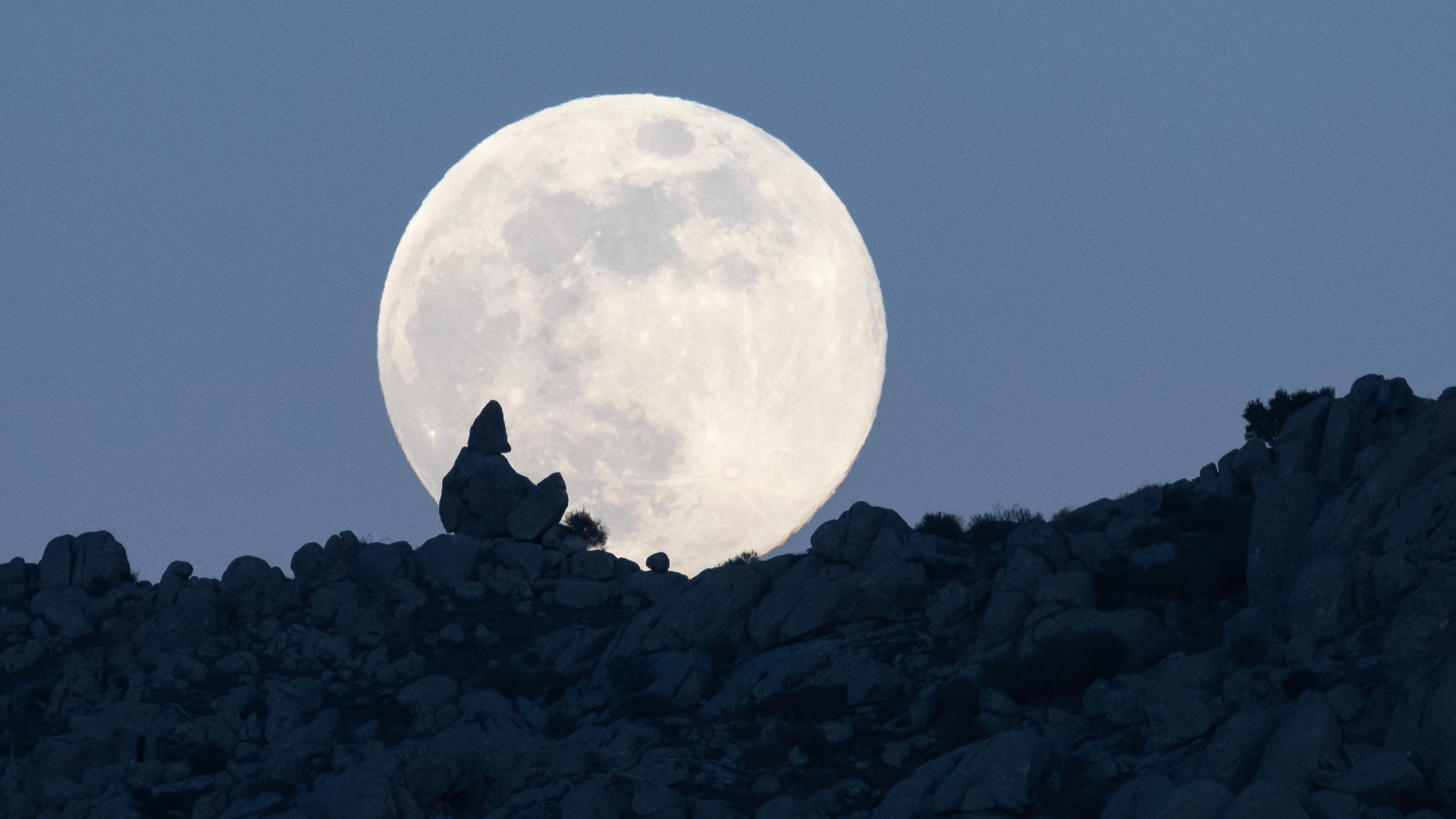 A full moon is photographed rising above a rocky hillside in the dark blue evening sky.
