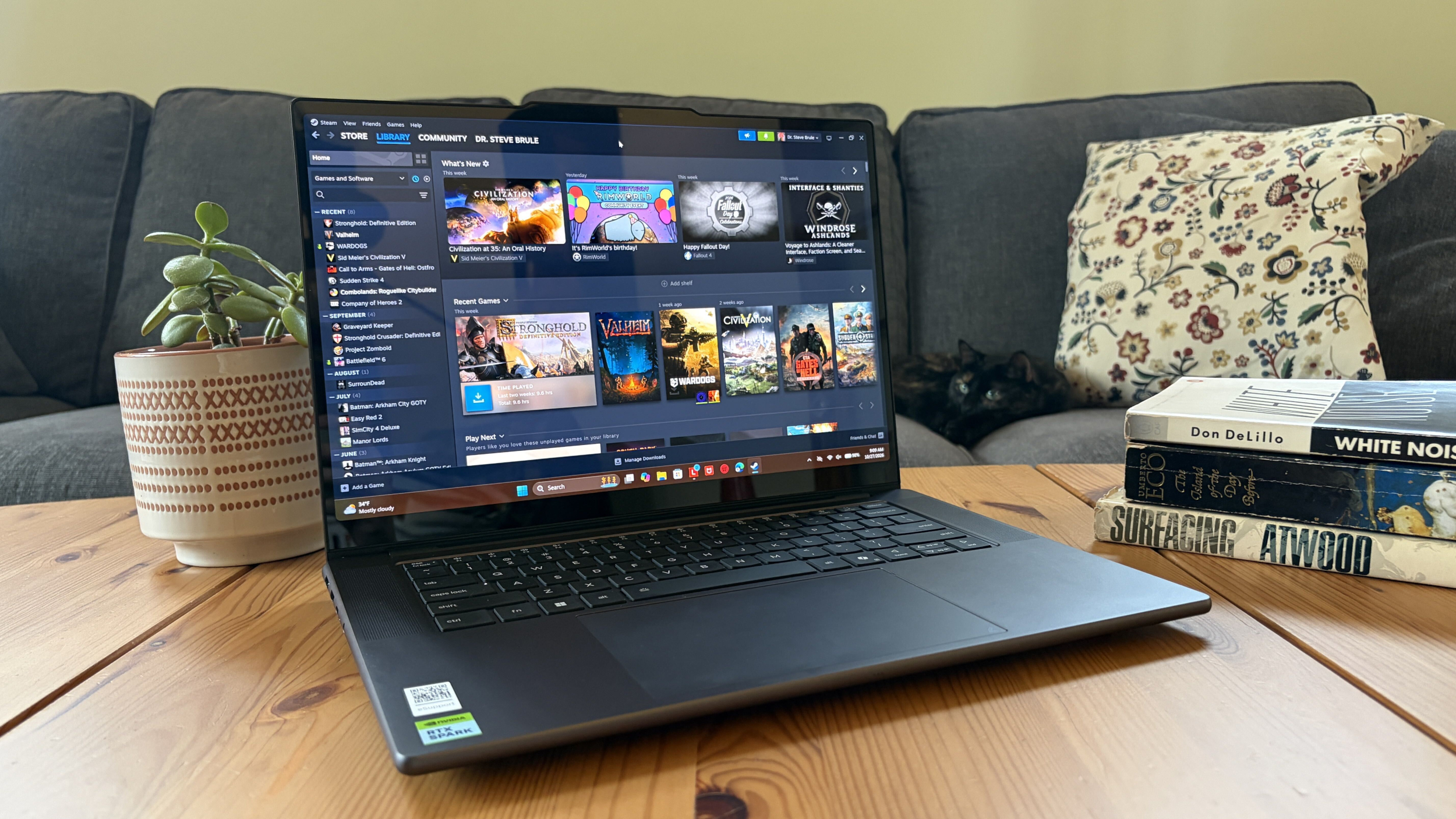 A Lenovo Pro 9n laptop showing a digital game library sits on a wooden coffee table, beside a potted plant and stacked books, with a patterned cushion on the sofa.