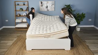 Two women maneuver the Saatva Classic mattress onto a bed frame in a bedroom.