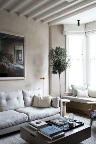 neutral living room with window seat, artwork, tree, pale grey sofa, coffee table, brass floor lamp