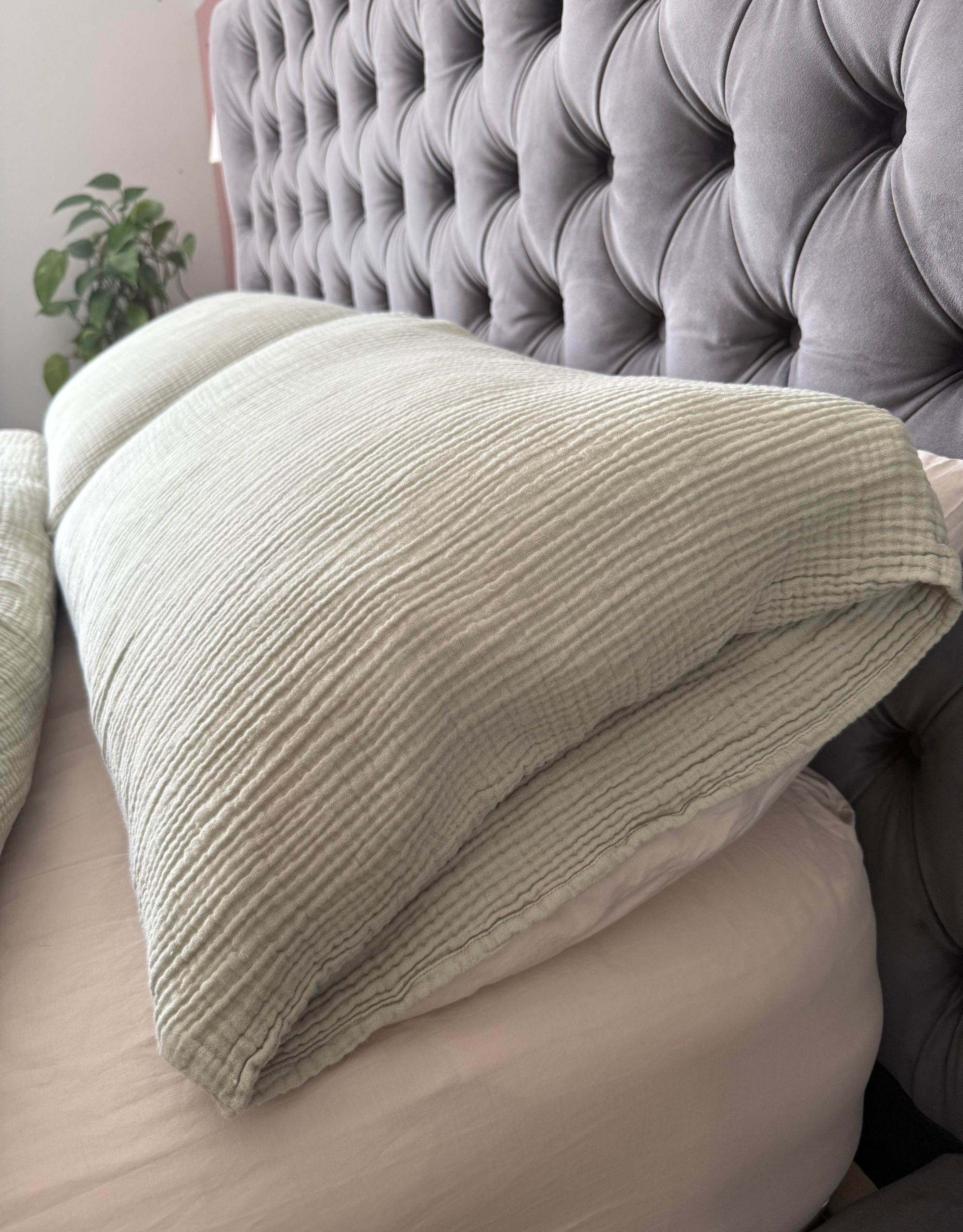 A close up of two light green muslin pillowcases with envelope openings on top of a beige sheet and by a dark gray-blue headboard