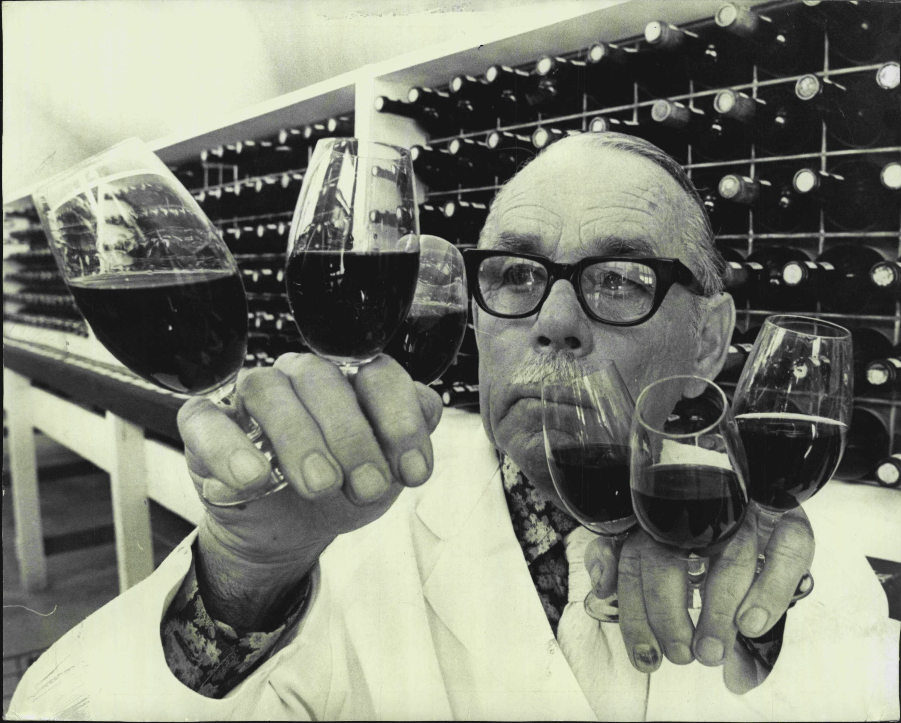 old man in glasses and lab coat holding six glasses of red wine, with wine barrels behind him