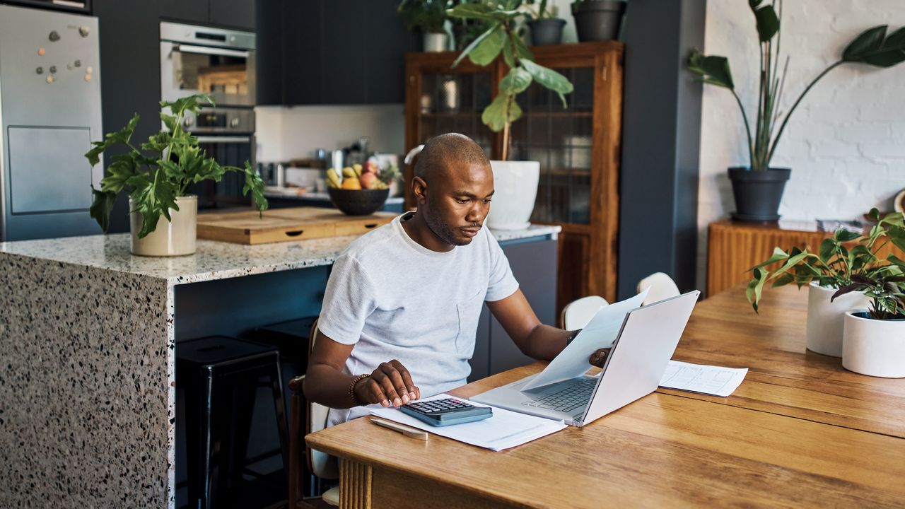 A man sits at his dining room table, looking at paperwork and using a calculator. 