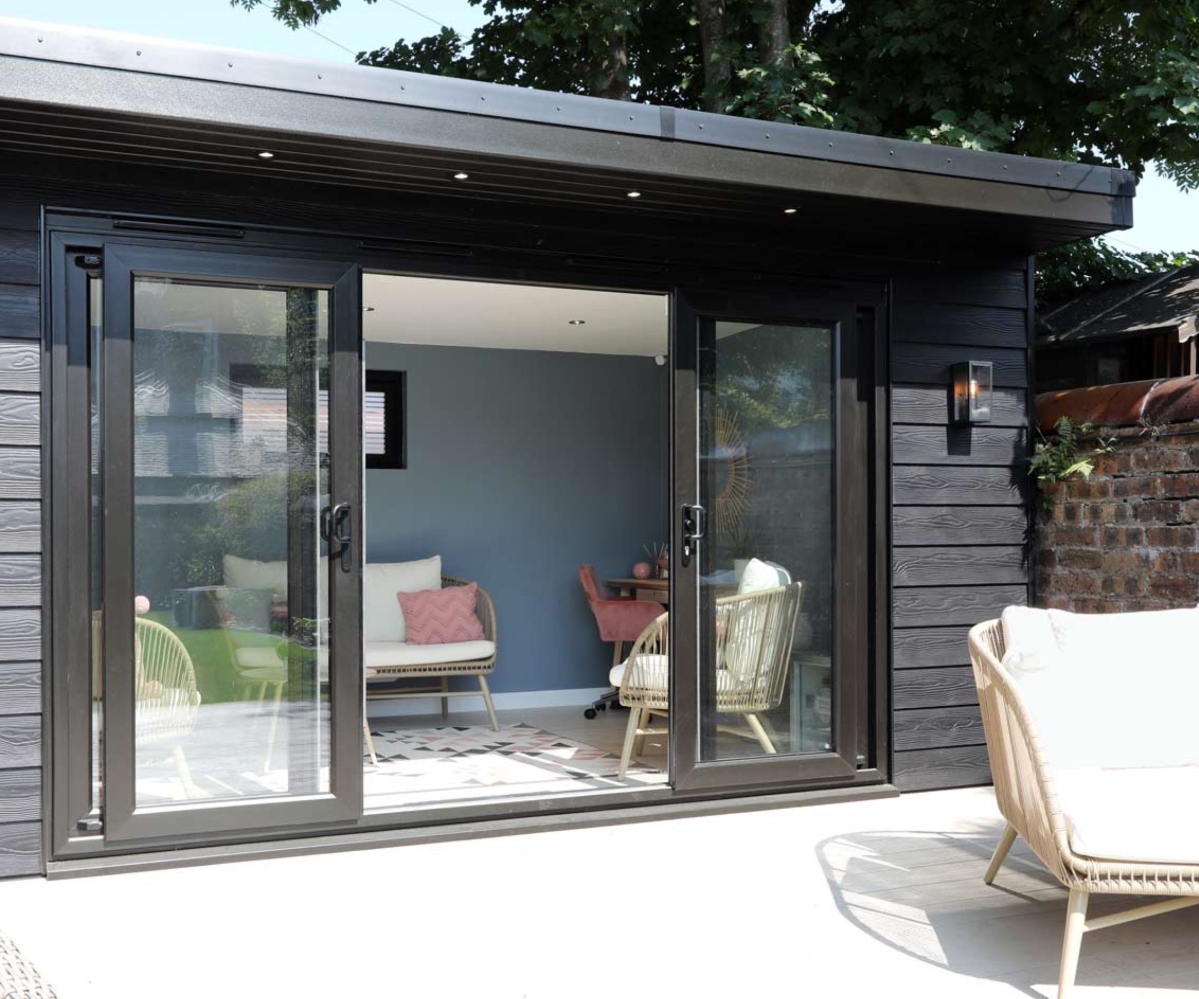 A charcoal timber-clad flat-roof garden room with full-width sliding glass doors open to reveal a blue-walled interior with rattan chairs and a geometric rug.