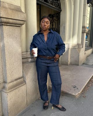 A woman wears dark blue jeans, black ballet flats, a denim top, and a brown belt.