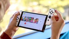 Nintendo Switch OLED held between two hands with one of the JoyCons being slid off