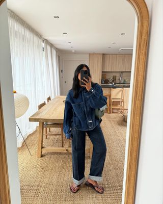 Monikh wearing a dark denim jacket and matching cuffed jeans with flip flops inside of her home.