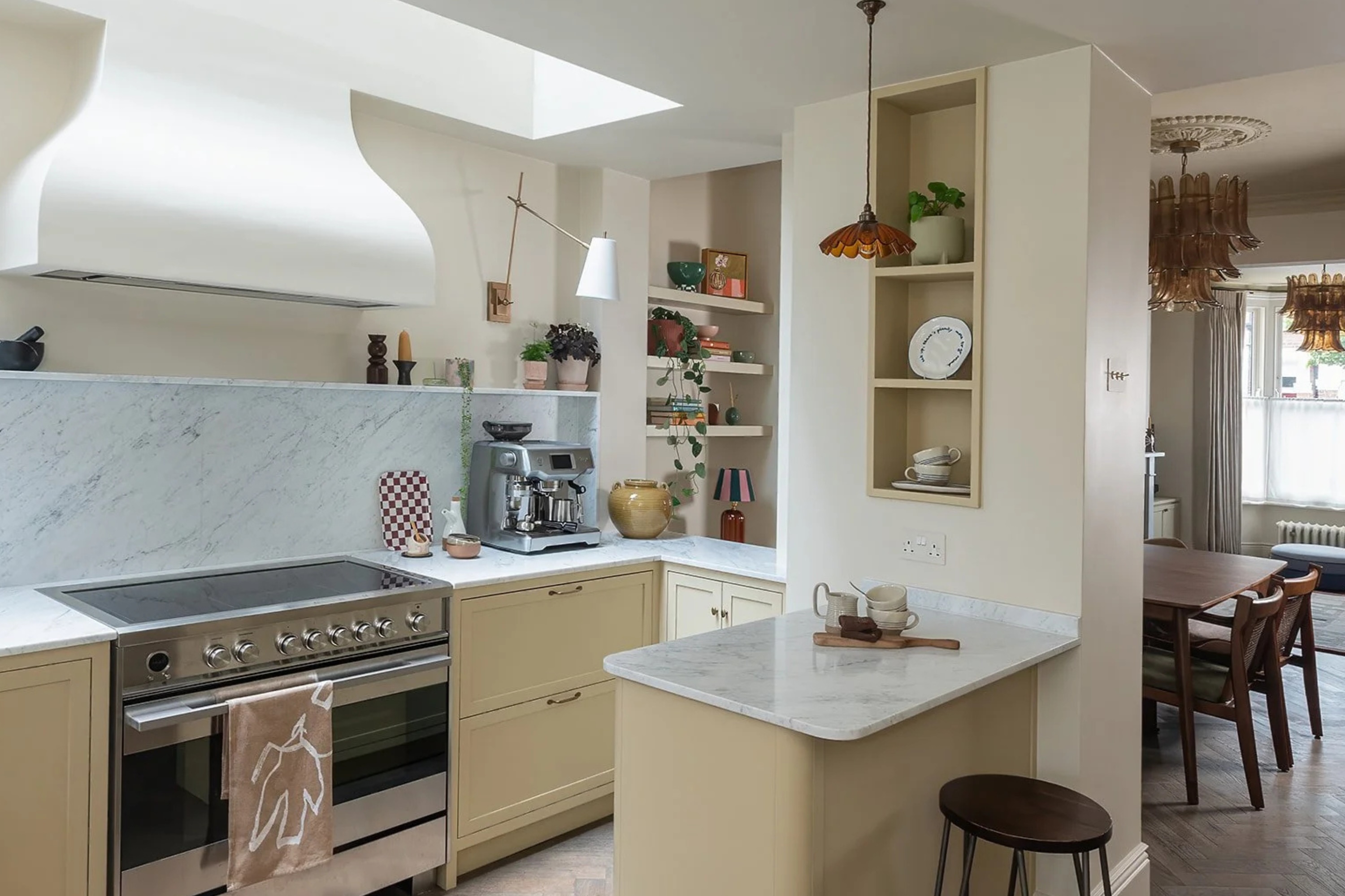 Small cream color kitchen with recessed open shelving, view through to dining room with glass chandeliers over a table