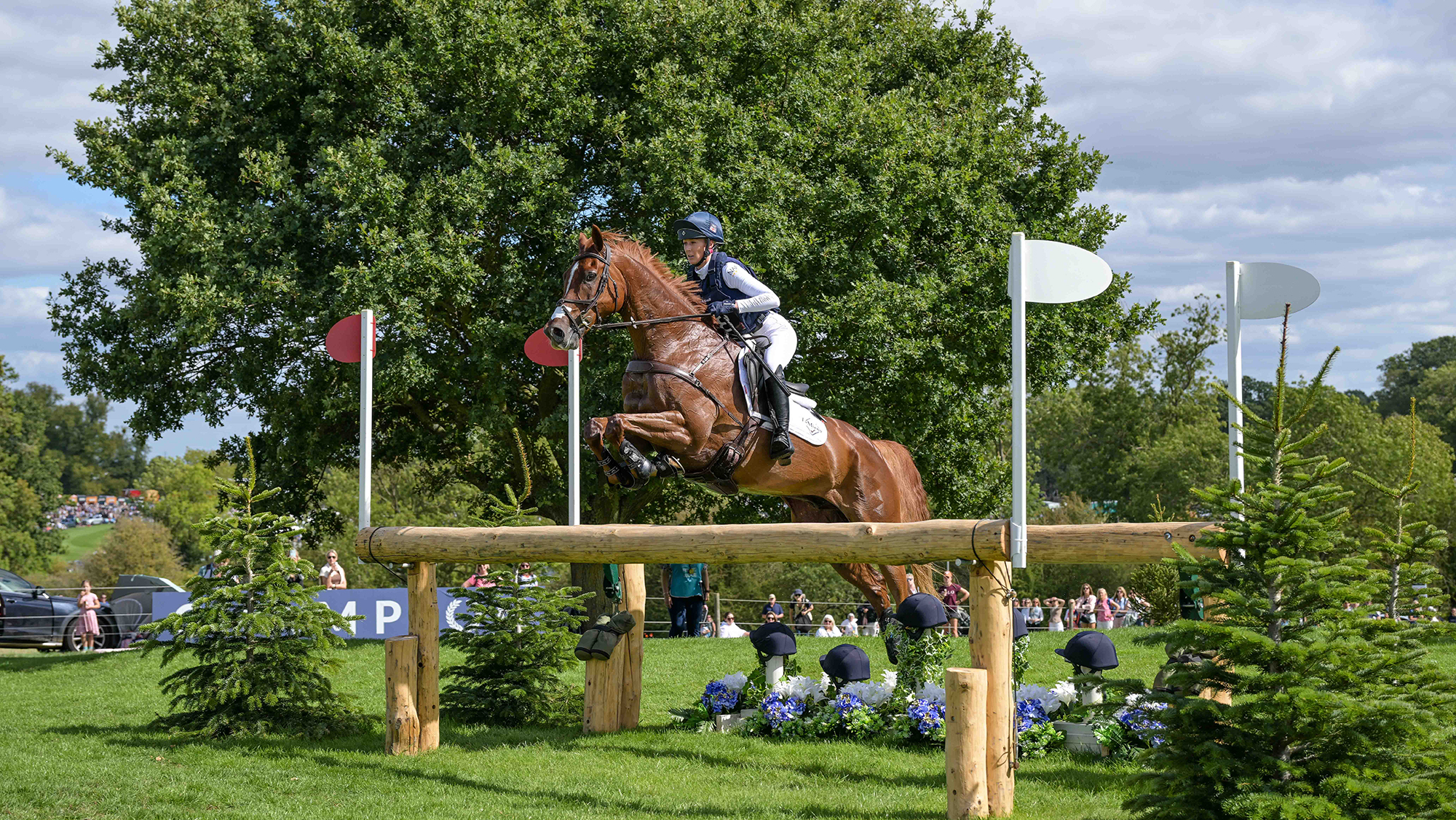 Yasmin Ingham, a rider in a white top with a blue body protector over it, rides chestnut horse Goliath Du Loir over a log fence at the 2026 Defender Burghley Horse Trials