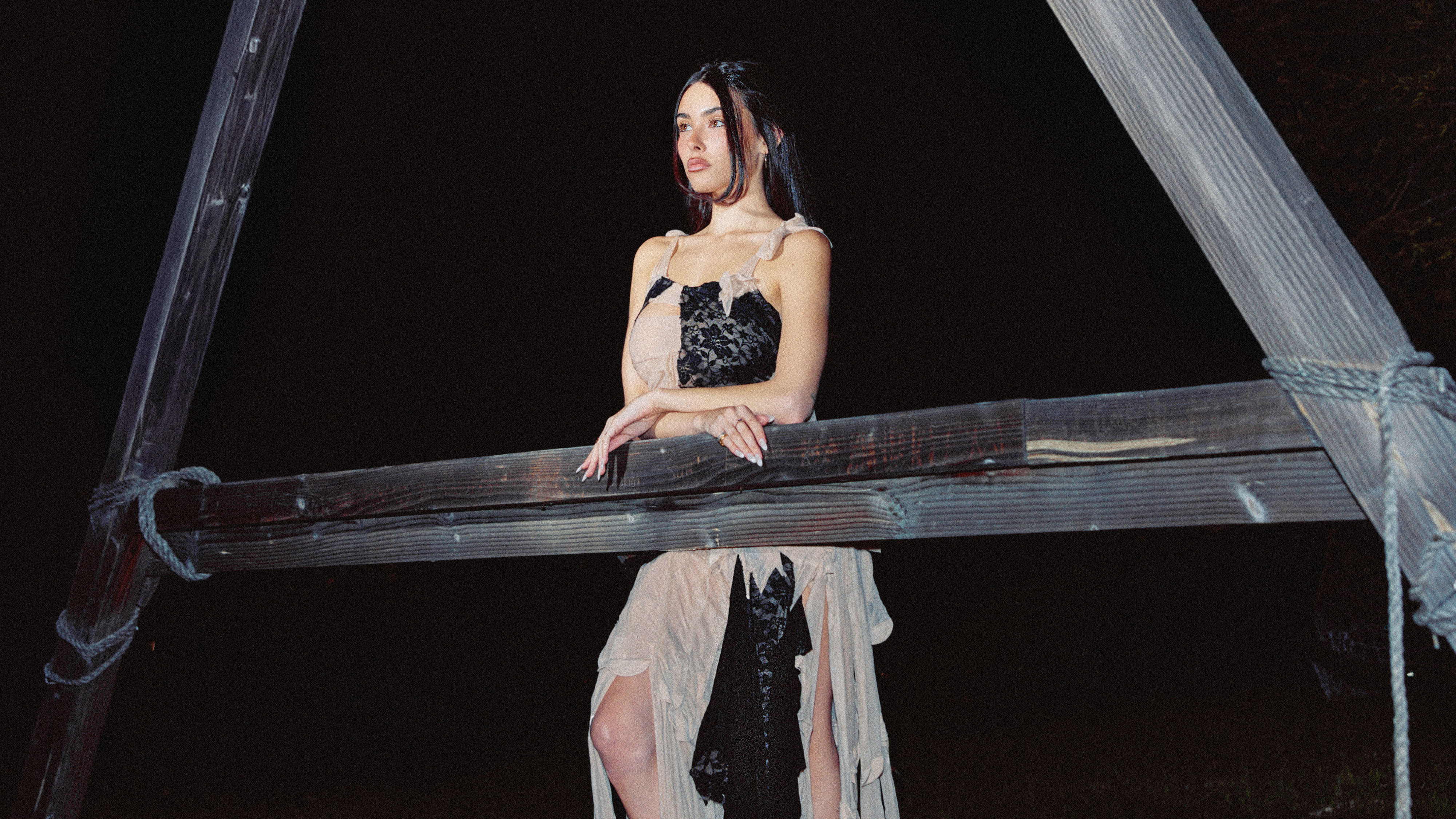 Image from Who What Wear's February cover story with Madison Beer. She is leaning on a wooden A-frame structure wearing a black and nude dress. 