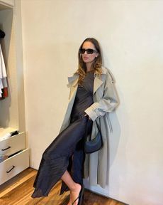 Pia Mance leans against wall wall wearing black slip dress and tan trench coat