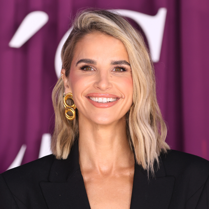 Vogue Williams close-up. She is smiling towarss the camera, wearing a black dress and gold earrings.