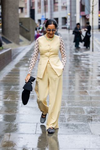 Zeena Shah mengenakan rompi kuning, kemeja panjang dengan cetakan logo, celana, tas hitam, kacamata hitam di luar 16Arlington selama London Fashion Week