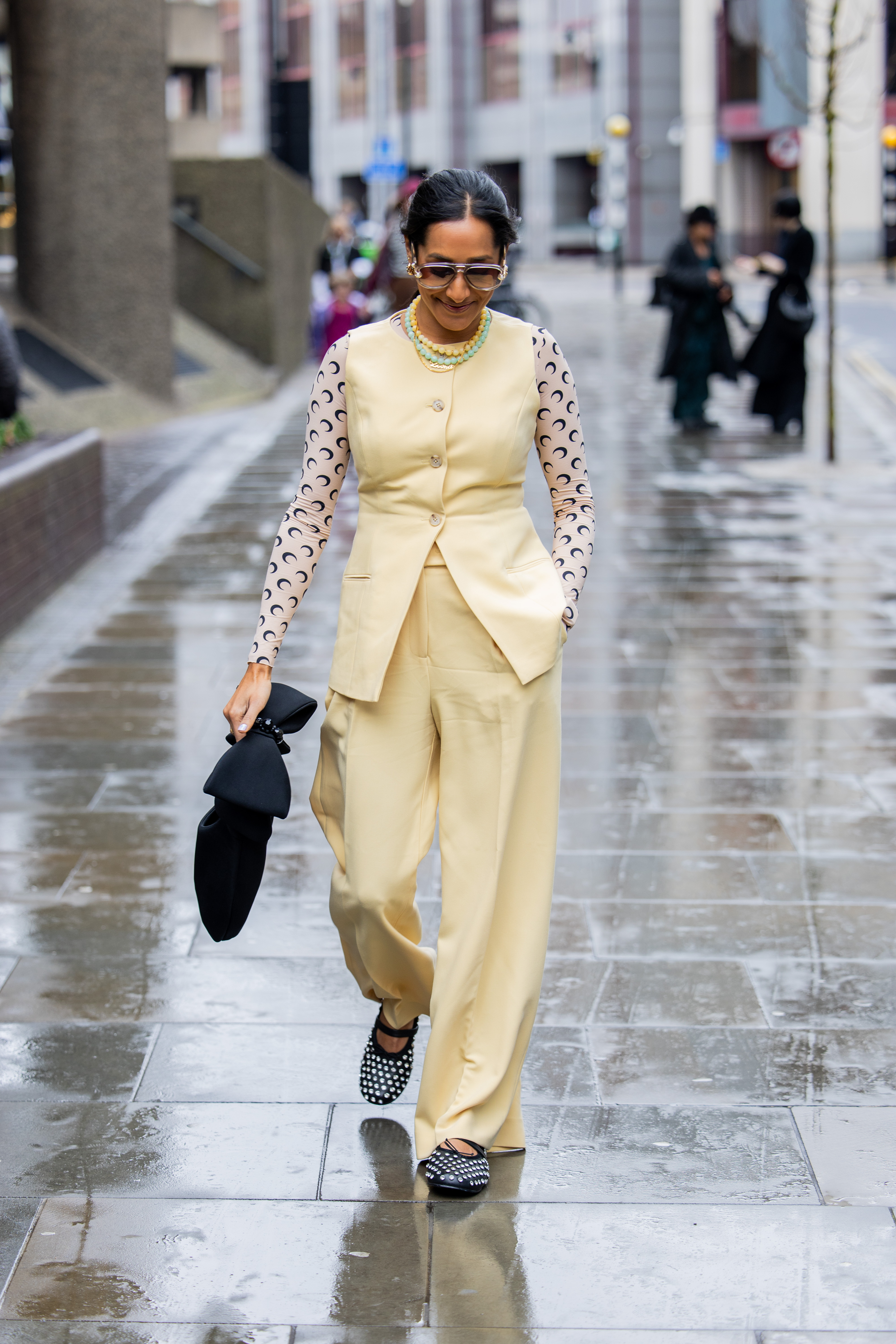 Zeena Shah wears yellow vest, long shirt with logo print, pants, black bag, sunglasses outside 16Arlington during London Fashion Week