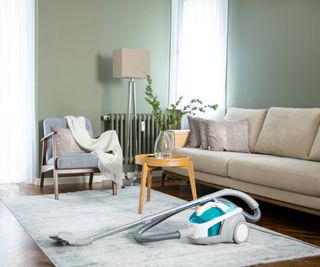 A blue and white vacuum cleaner on a cream living room rud, beside a cream couch and a small yellow side table