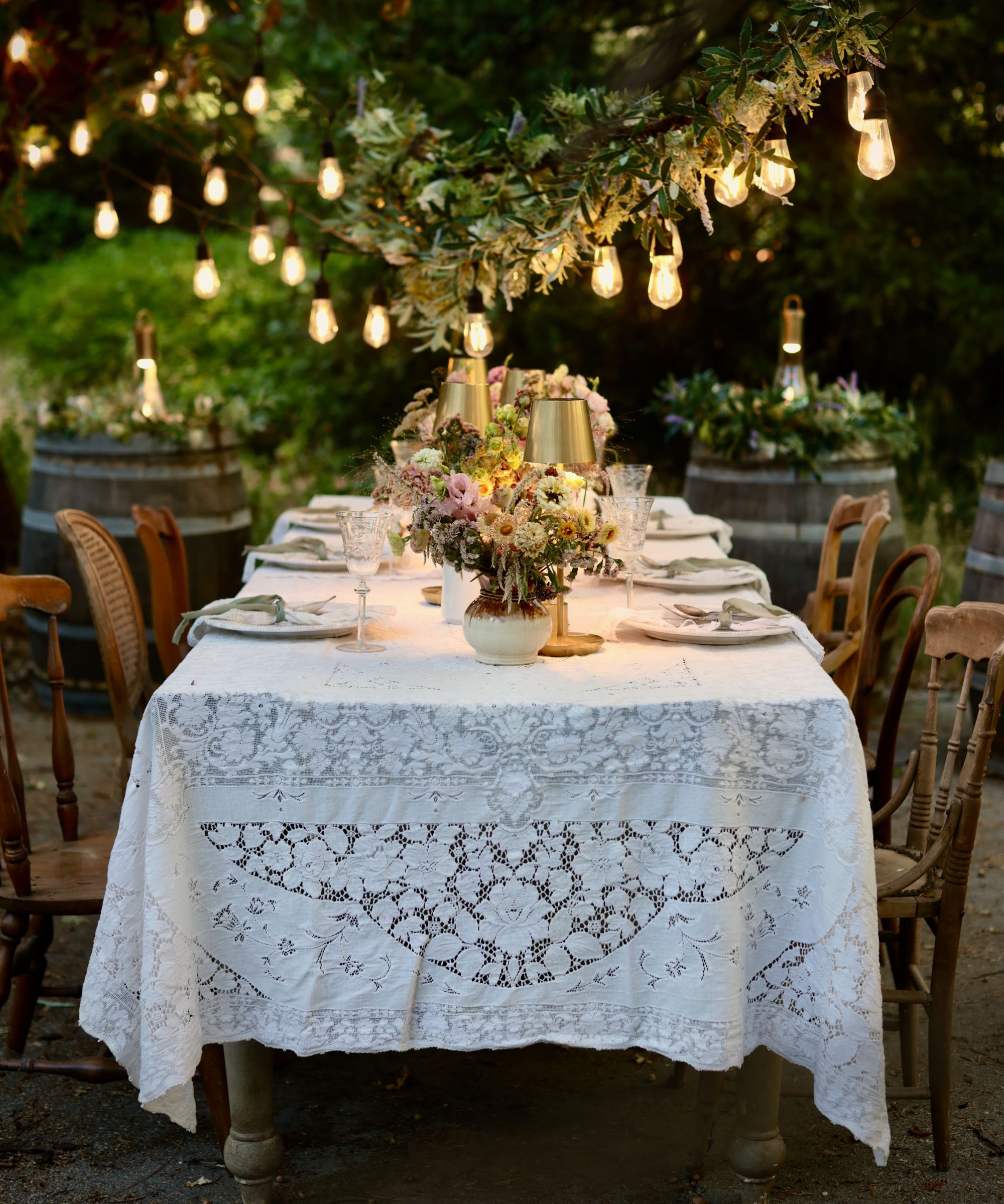 outdoor dining table with festoon lighting, a lace tablecloth, flowers and lamps