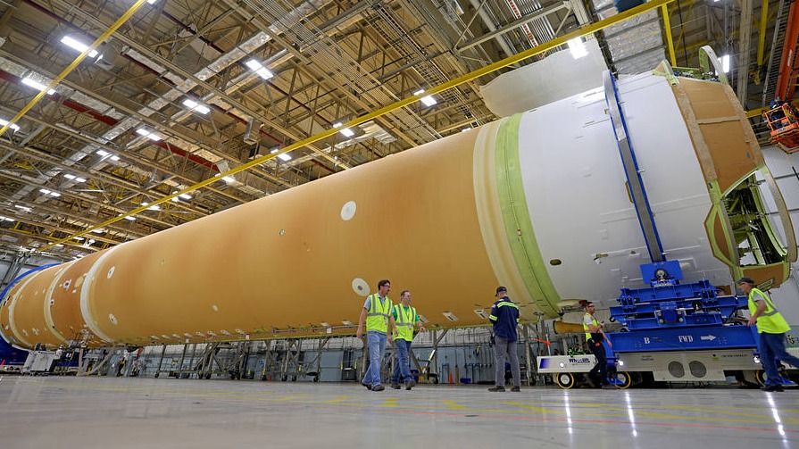 orange rocket stage lies on its side in a big hangar. white stripes mark the edge of the rocket and a machine of some sort sits in front of it. technicians walk in front of the rocket with yellow safety vests