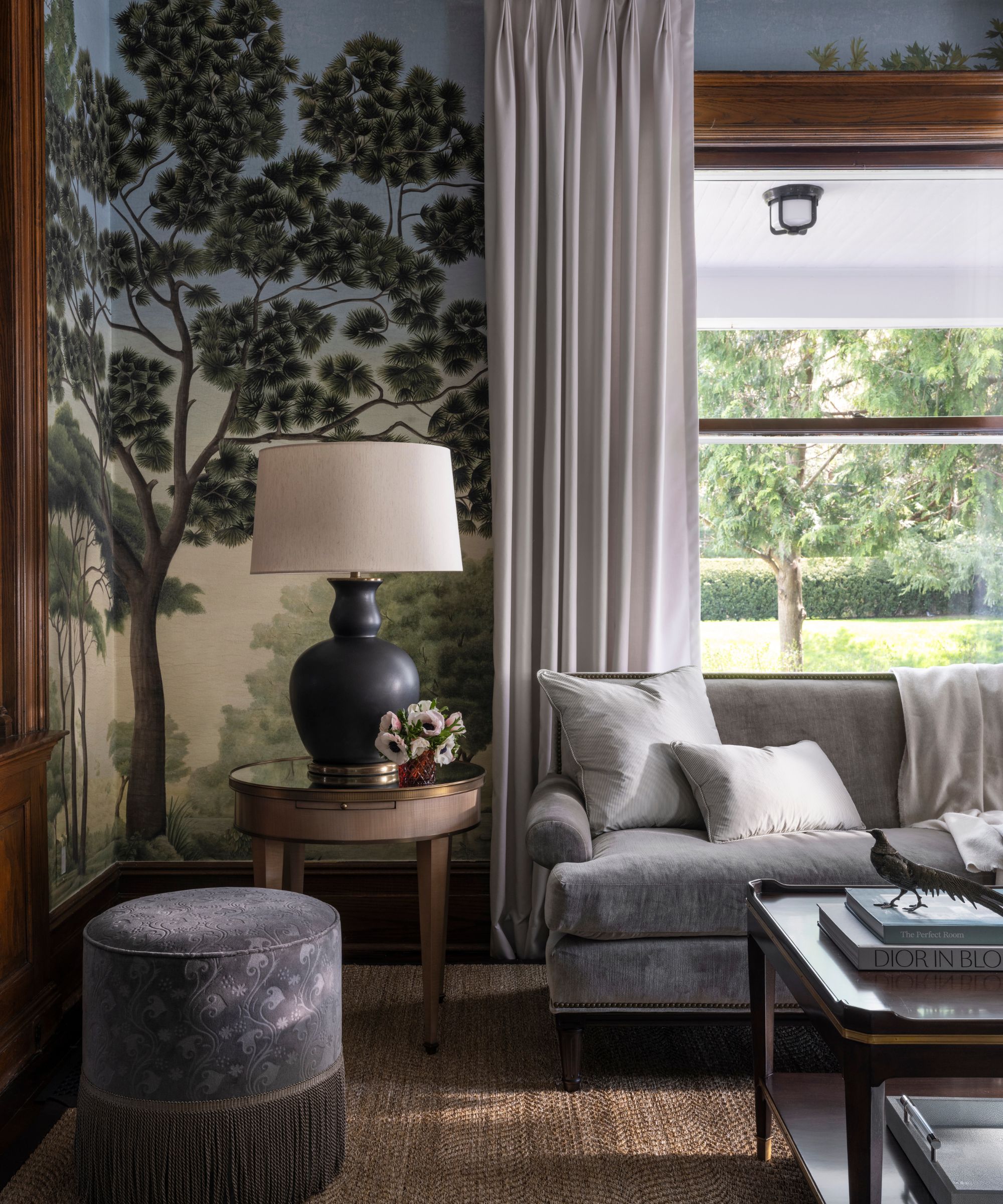 Living room with mural wallpaper, grey velvet sofa, a side table with a large lamp on top, a large window overlooking a garden, a jute rug, fringed ottoman, and a lacquer coffee table.