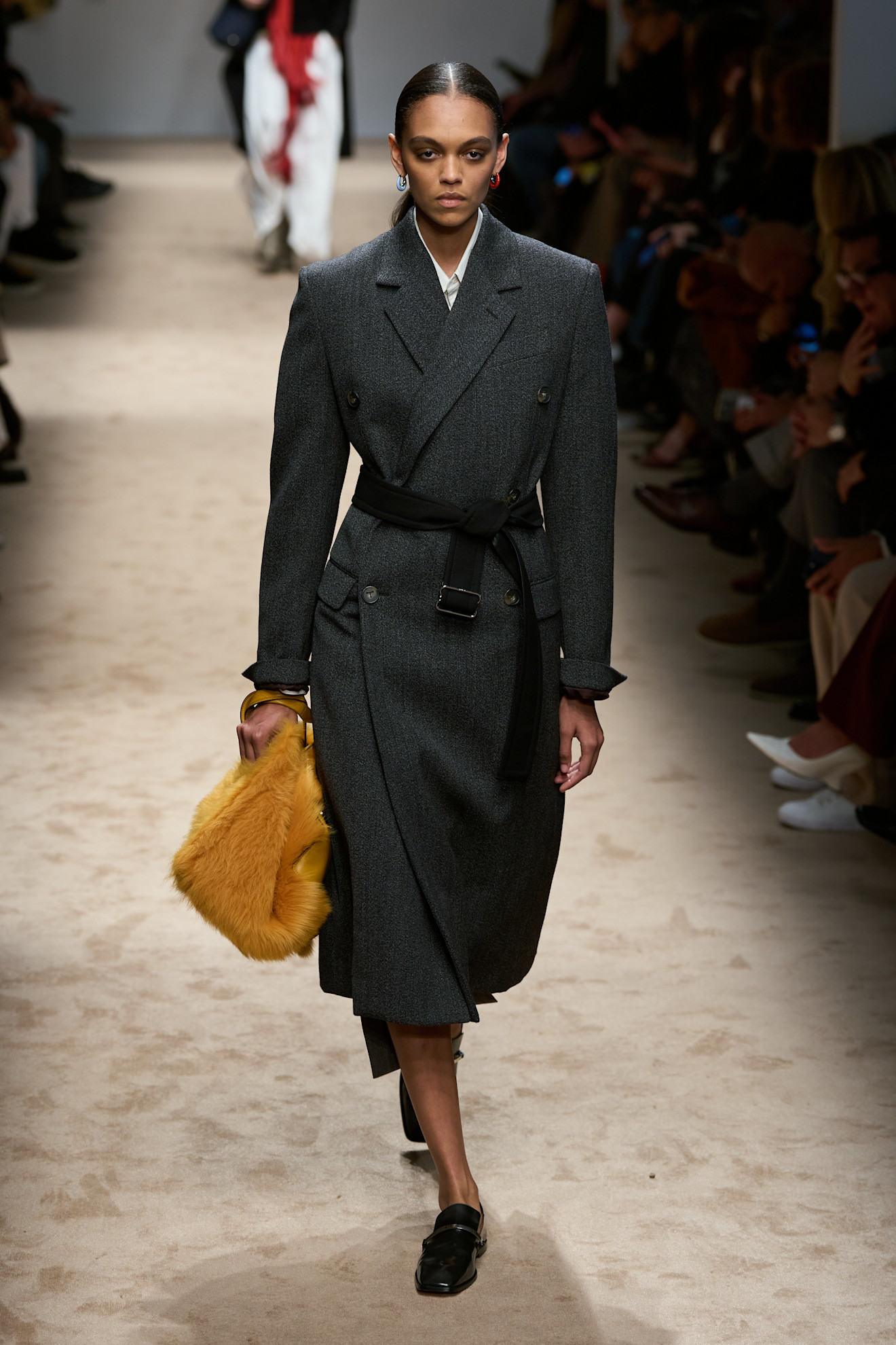 Fur accessory trend is shown in Tod's fall-winter 2025 collection in a photo of a model walking down the runway wearing a tailored gray coat over a white shirt and gray skirt styled with a black belt on top, black loafers, and a yellow fur handbag