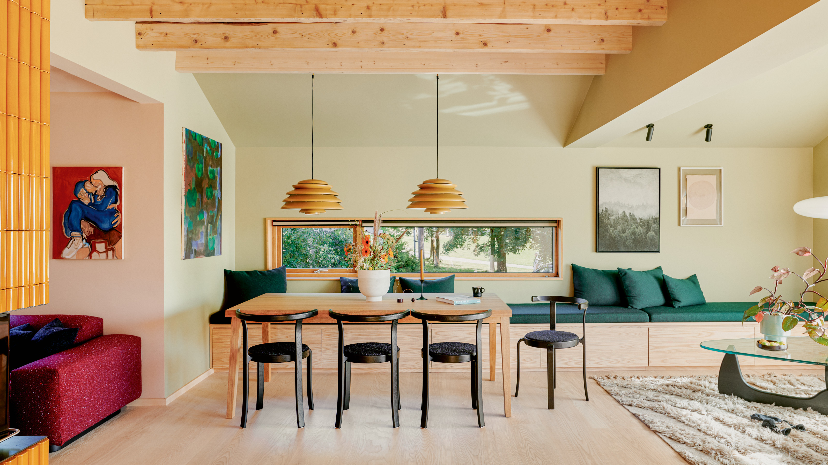 Dining area of an open-plan space with built-in dining bench with storage, light wood table, black chair, double pendant lights, exposed wood ceiling and rafters