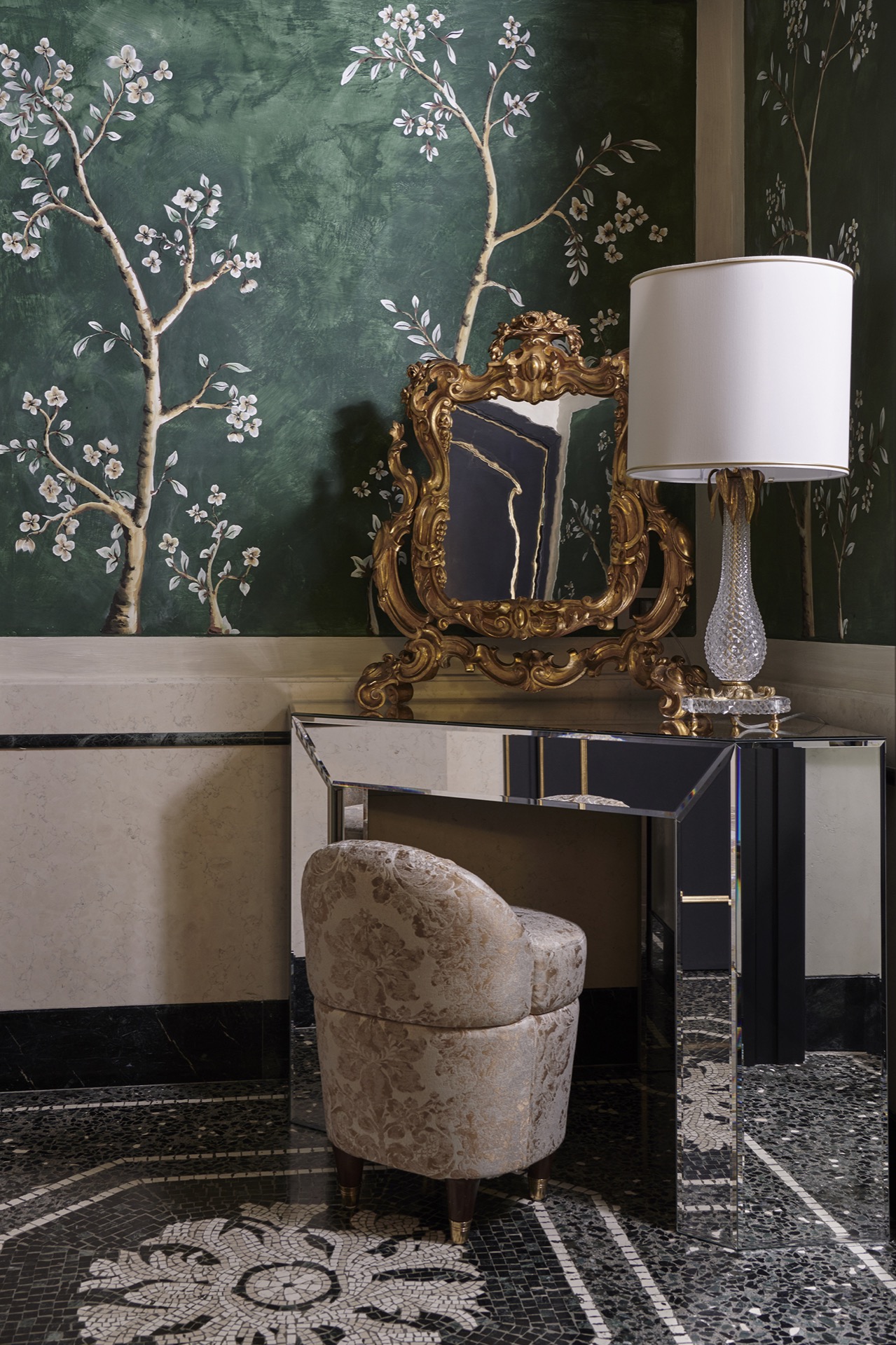 A transitional hotel room juxtaposes classical mosaiced floors with a floral motif with a mirrored, geometrical desk, a velvety chair, Murano glass-carved table lamps, golden mirrors, and mural-clad walls.