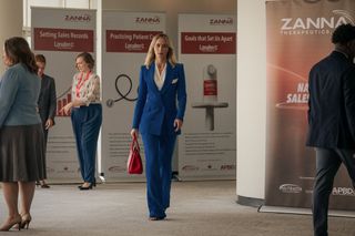 Emily Blunt in Painkiller walking past posters for Zanna, the pharmaceutical company