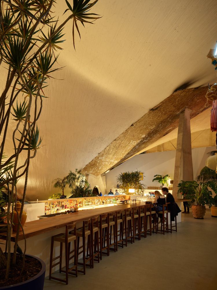 A golden-lit restaurant housed inside a modernist building with exposed rock surfaces and contemporary wooden design boasts a long wooden bar counter with matching stools in dark wood and ochre fabrics.