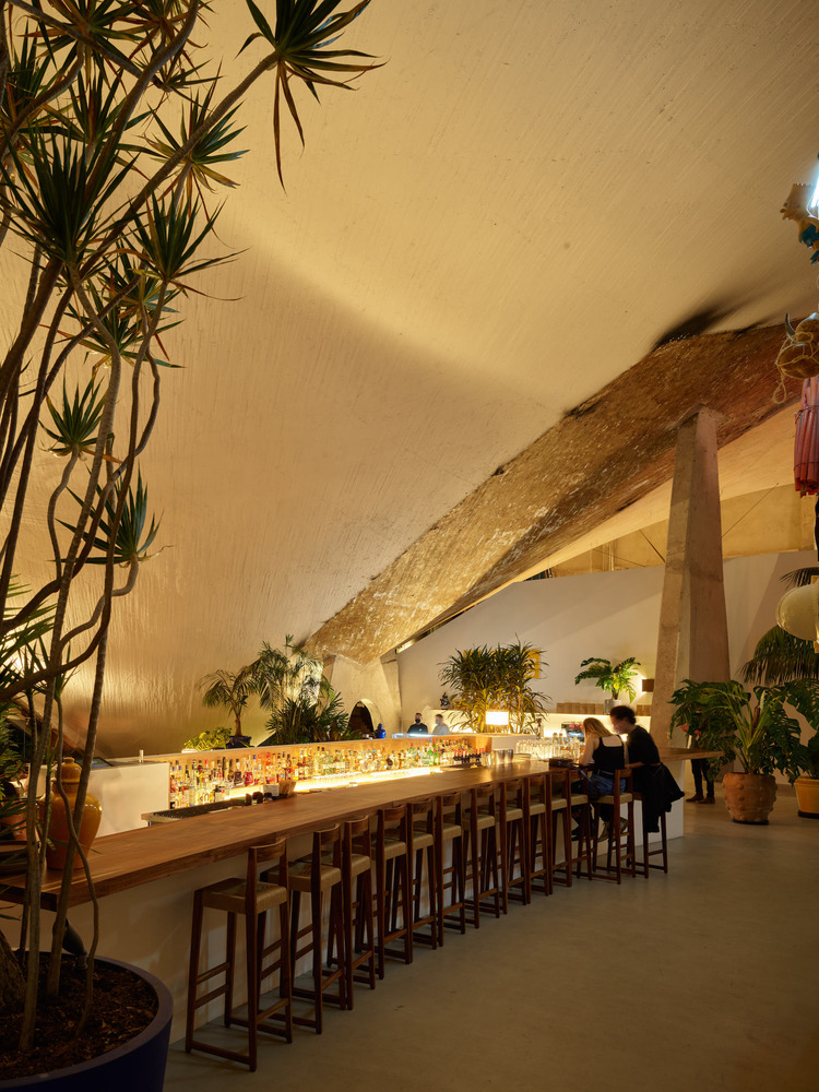 A golden-lit restaurant housed inside a modernist building with exposed rock surfaces and contemporary wooden design boasts a long wooden bar counter with matching stools in dark wood and ochre fabrics.