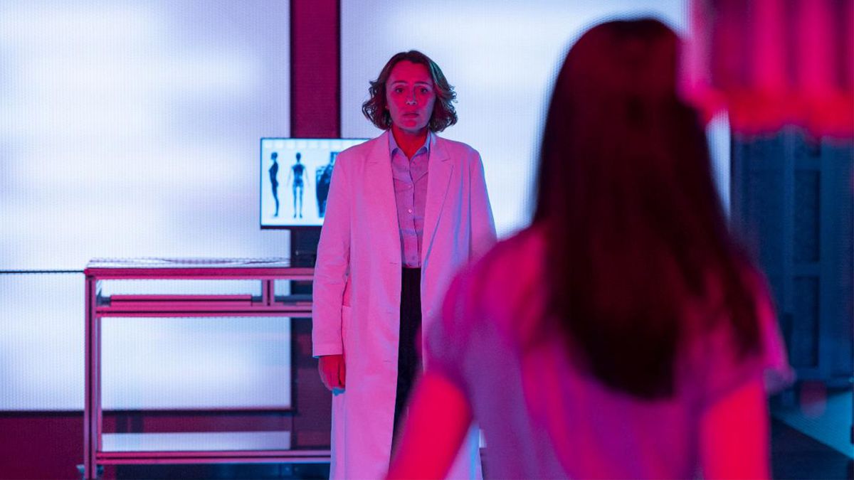 A woman wears a lab coat in a hospital room with an x-ray body scan displayed behind her