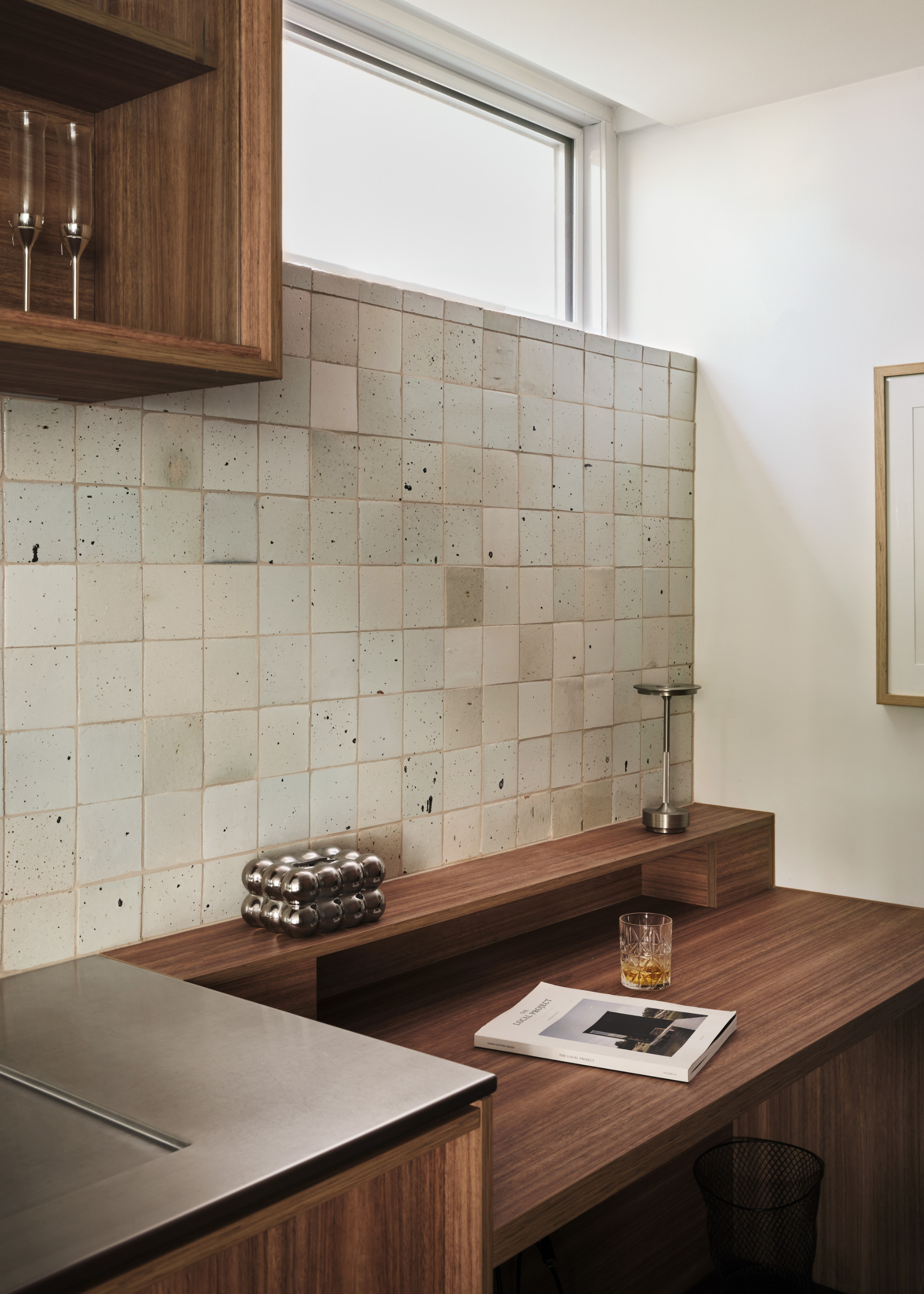 A wood extension with a bubbled chrome coaster, a glass of liquor and a book near a stone tile backsplash