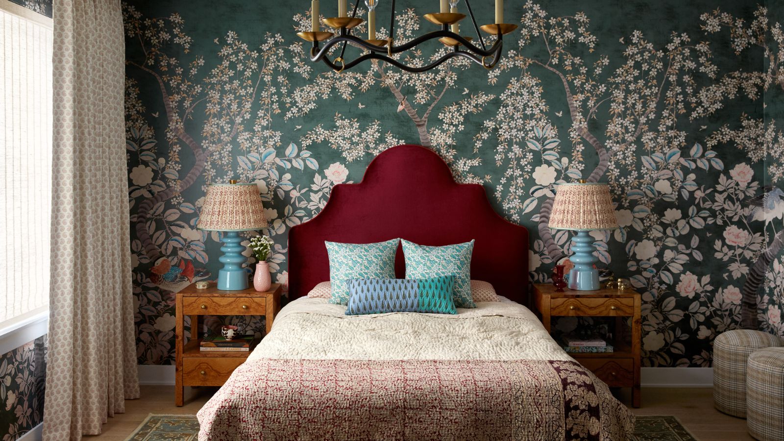 A bedroom with teal floral wallpaper, a burgundy headboard, blue and burgundy bedding, and two wooden nightstands with blue lamps. 