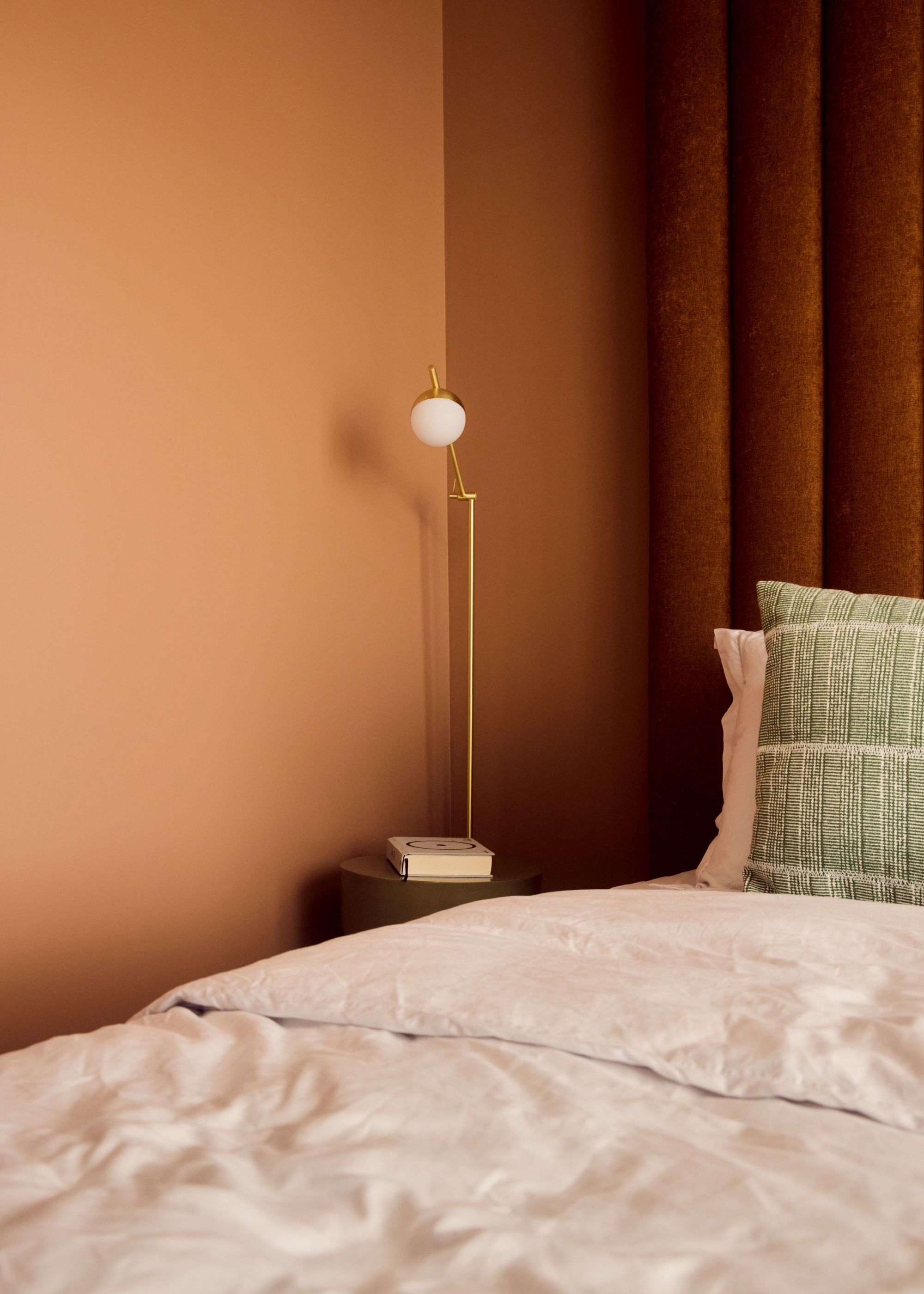 A corner of a bedroom painted terracotta with matching curtains, and linen bedding with a sage green cushion.