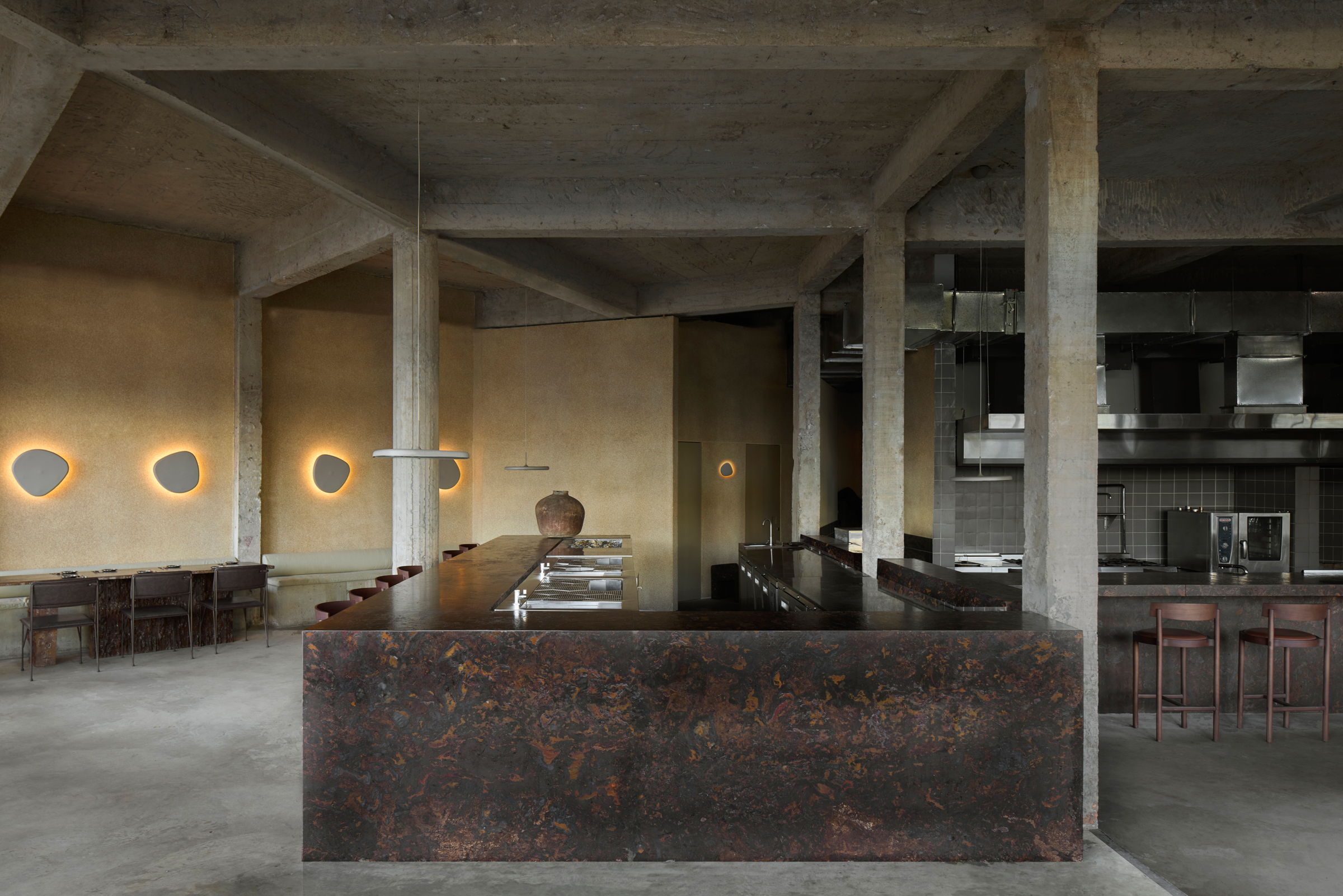 A brutalist building houses a sleek, almost utopian restaurant crafted from chocolatey slabs of stone, sheeny chrome, and exposed cement columns, with organically shaped, back-lit sconces resembling rocks.