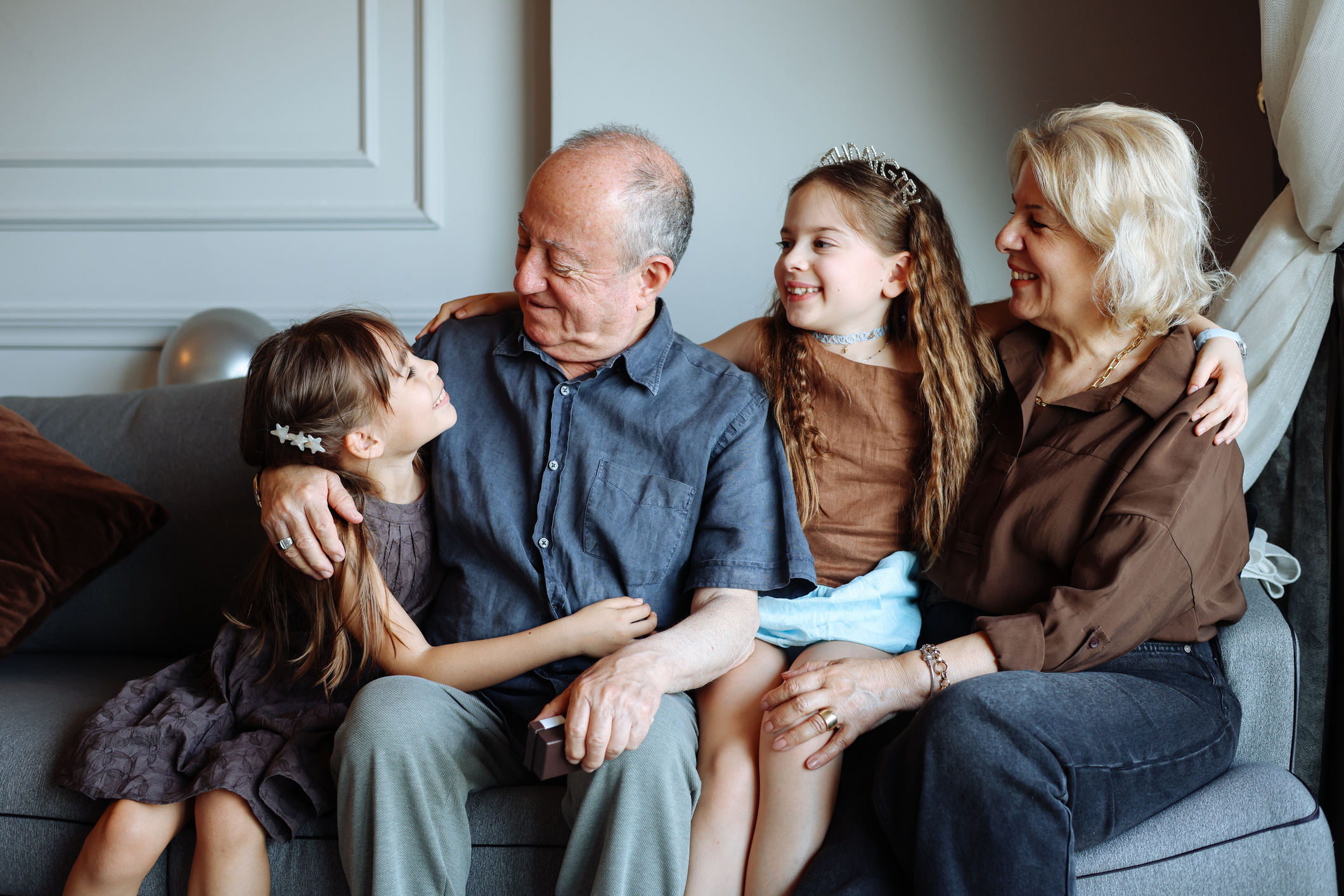 A heartwarming indoor lifestyle photograph showcasing a senior couple sharing a tender moment with their granddaughters on a cozy gray couch. 