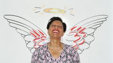An older woman leans against a wall that has angel wings and a halo drawn on it. She is smiling and her eyes are closed.