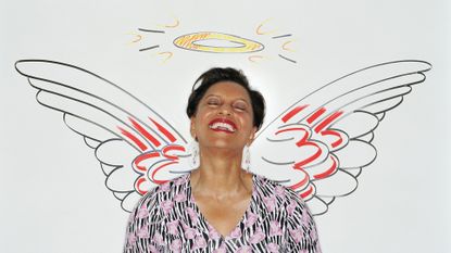 An older woman leans against a wall that has angel wings and a halo drawn on it. She is smiling and her eyes are closed.