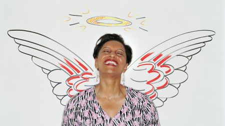 An older woman leans against a wall that has angel wings and a halo drawn on it. She is smiling and her eyes are closed.