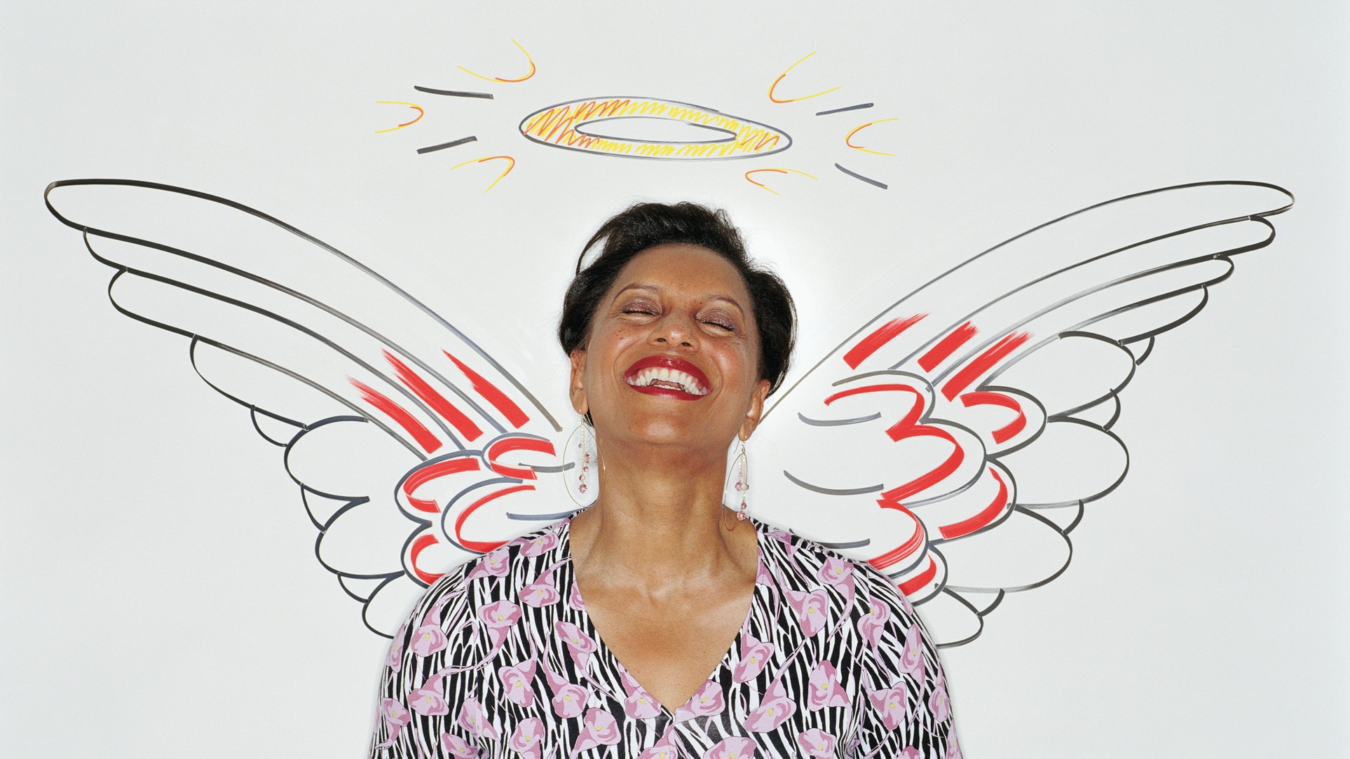 An older woman leans against a wall that has angel wings and a halo drawn on it. She is smiling and her eyes are closed.