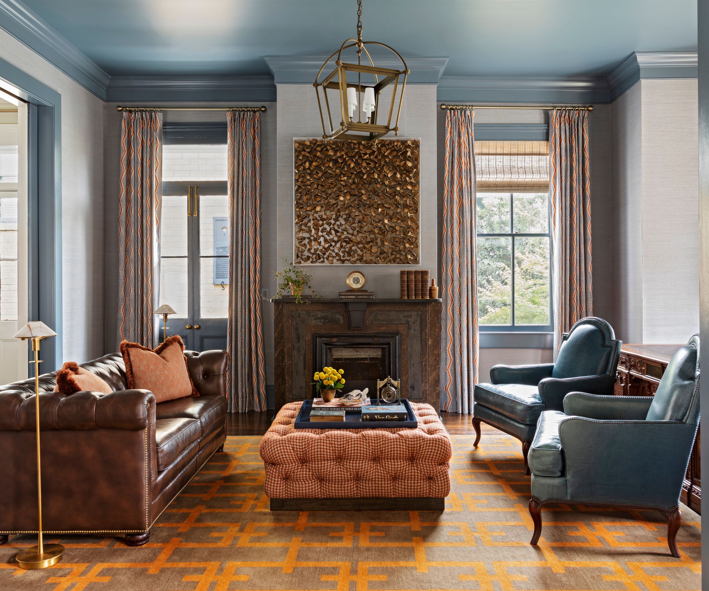 Living room with blue ceiling, gold antique pendant light, fireplace, upholstered ottoman, leather sofa, and blue leather armchairs with orange 1970s style rug