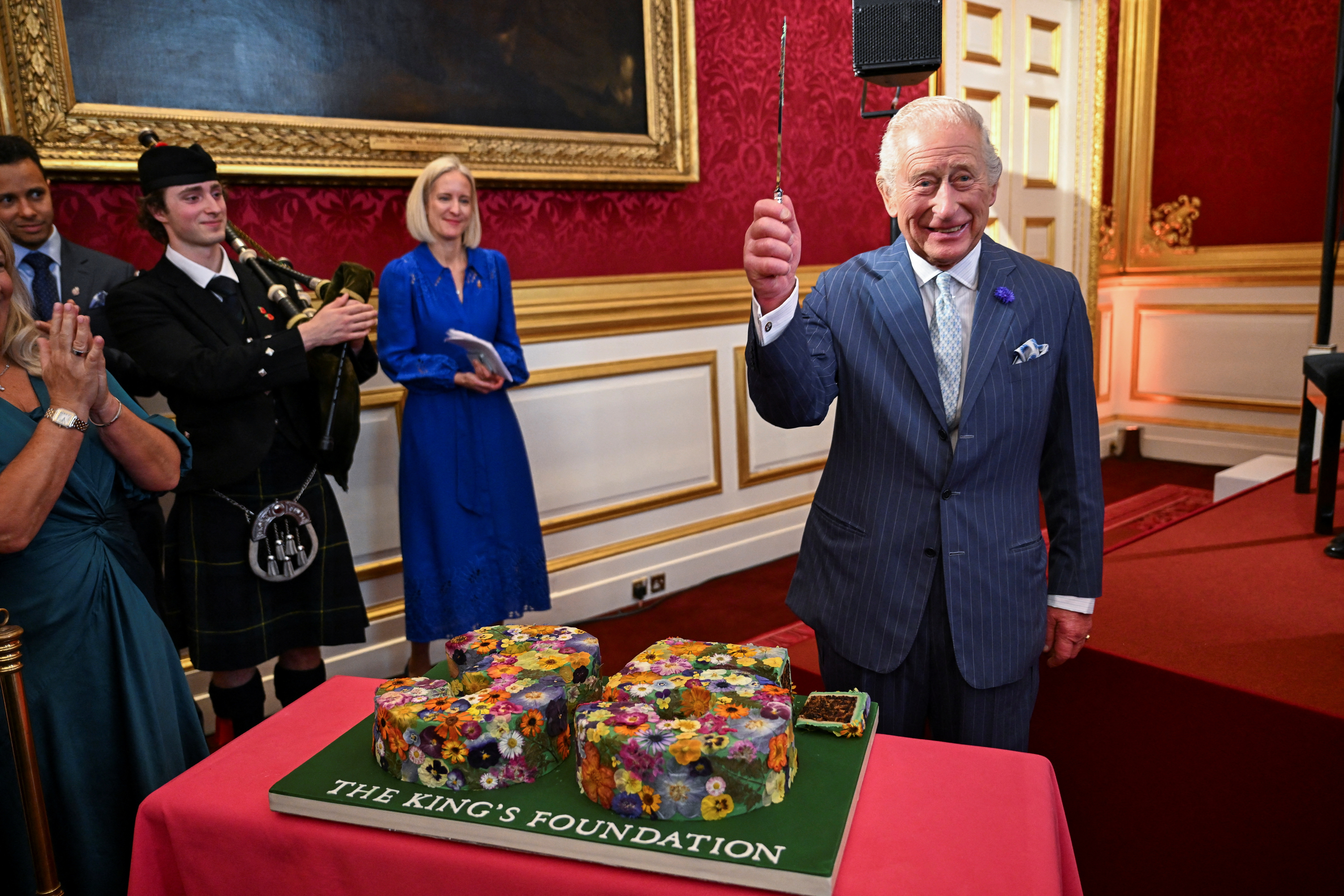 King Charles wearing a blue suit holding a cake knife in front of a cake reading The King&#039;s Foundation