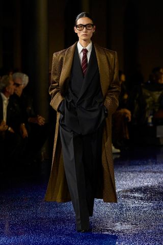 A model walking at the Saint Laurent S/S 25 runway show during Paris Fashion Week wearing a suit, shirt, and tie with a brown silk coat on top.