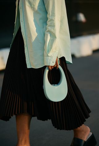 a close-up photo of a woman wearing a blue shirt with a black pleated skirt and a coperni designer bag