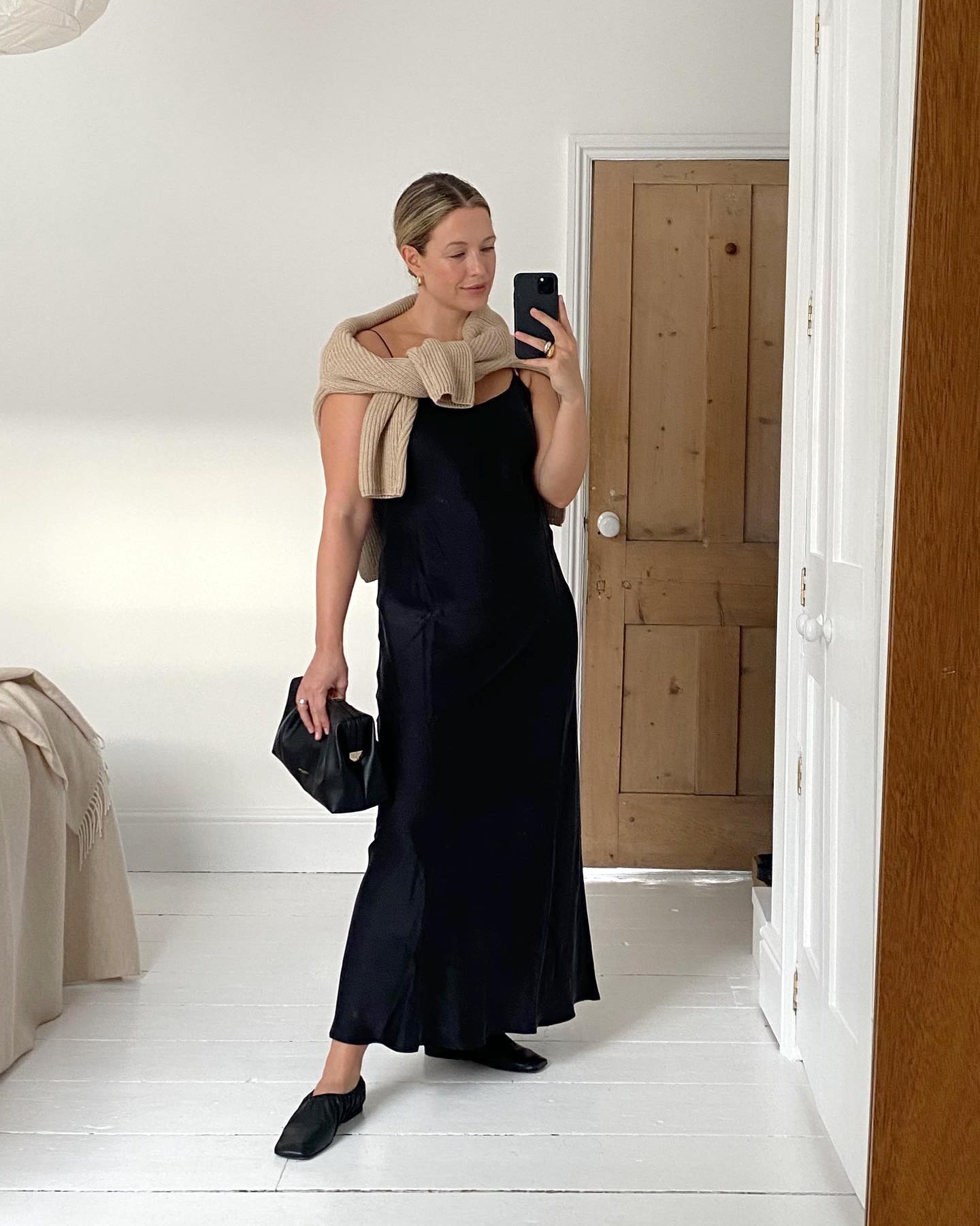 British influencer Alexis Foreman poses for a mirror selfie in a minimalistic bedroom with white wood floors wearing a neutral tan sweater tied over her shoulders, a black slip dress, a black clutch bag, and black leather flats.