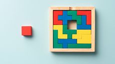 A wooden puzzle made up of colored blocks has missing piece in the center: a red square that literally sits outside the box.