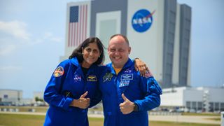 two astronauts in flight suits giving thumbs up outside. far behind them is a square building with a flag and nasa logo