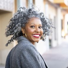 Black woman with grey curly hair and red lipstick wears grey coat and gold hoop earrings