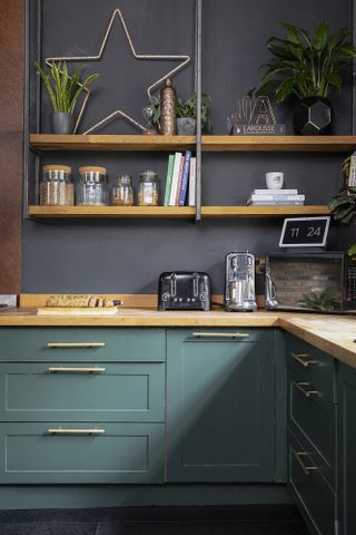 Green Shaker-style kitchen against black wall, with wood open shelving and brass handles