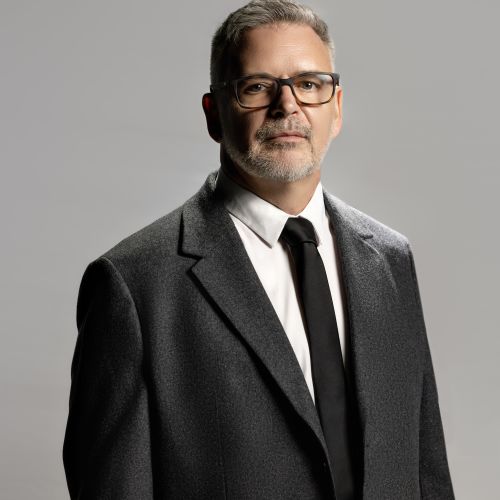 Photograph of person in black and white suit and dark glasses, with gray hair and short facial hair.